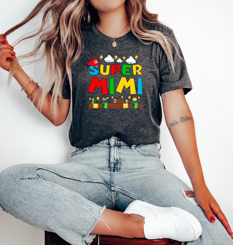 May include: A gray t-shirt with a graphic print of the text "SUPER MIMI" in colorful letters, inspired by the Super Mario Bros. video game. The graphic also includes a red cap, clouds, coins, and a green pipe.