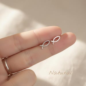 Op de afbeelding: Een paar kleine, zilverkleurige oorstekers in de vorm van een vis. De oorbellen worden op de vinger van een persoon getoond. Het woord "Natural" staat in cursief schrift in de rechter benedenhoek.