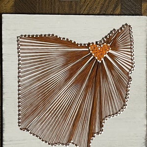 May include: String art of the state of Ohio with a heart in the center. The outline of the state is made with white string and the heart is made with orange string. The background is a light wood color.