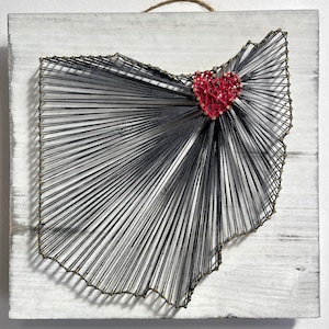 Puede incluir: Una pieza de arte de cuerda que representa el contorno del estado de Ohio con un corazón rojo en el centro. El arte se crea con cuerda negra envuelta alrededor de clavos martillados en una tabla de madera blanca.