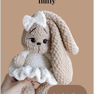 May include: A handmade, fluffy crochet bunny in beige and white. The bunny has long ears, a white bow, and a white dress. The text "Patroon: Little bunny fluffy" is at the top, and "Juudstudios" is at the bottom.