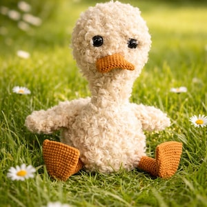 Op de afbeelding: Een handgemaakt eendenspeelgoed met een crèmekleurige, krullende textuur. De eend heeft een bruine snavel, poten en zwarte ogen. Het zit in groen gras met witte madeliefjes.