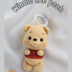 Op de afbeelding: Een gehaakte Winnie the Pooh sleutelhanger in beige met een rood shirt. De sleutelhanger heeft een witte ring en de tekst "Winnie the Pooh" boven de beer. Een honingpot en lepel staan in de linker benedenhoek.