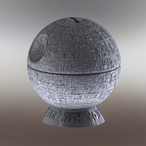 May include: A silver Death Star money box with a slot on top for coins. The base of the money box has the words "Be With You" printed on it.