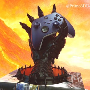 May include: A black, claw-like controller stand holding a blue Xbox controller. The stand is placed on a book with a fiery orange and yellow background. The stand has a spiked base and is branded with the text "@Primo3DDesign".