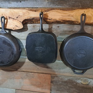 May include: Three cast iron skillets hanging on a wooden rack. The skillets are black and have a rustic look. The rack is made of natural wood with a live edge.