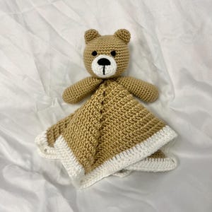 May include: A handmade crochet bear security blanket. The bear has a tan body, black and white facial features, and small ears. The blanket portion is tan with a white border.