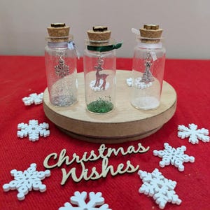 May include: Three clear glass bottles with cork stoppers, each containing a small metal charm: a snowflake, a deer, and a Christmas tree. The bottles are filled with glitter and tied with ribbons, displayed with wooden snowflake cutouts and the text Christmas Wishes.
