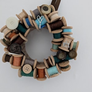 May include: A wreath made of wooden spools of thread, decorated with buttons, a sewing machine charm, and a pair of scissors.