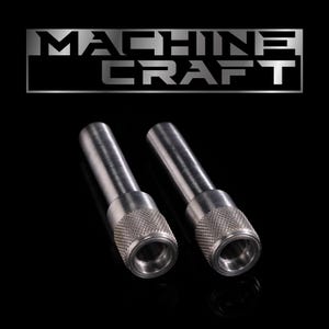 Puede incluir: Dos objetos metálicos plateados con un agarre moleteado en la base. Los objetos son cilíndricos con un centro hueco. El texto "MACHINE CRAFT" se muestra en plata, con una fuente en bloque sobre los objetos.