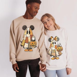 May include: Beige and off-white sweatshirts featuring a Halloween-themed Mickey and Minnie Mouse design. The characters are dressed as ghosts in patchwork costumes, holding jack-o'-lanterns. The sweatshirts are perfect for couples or friends celebrating the holiday.