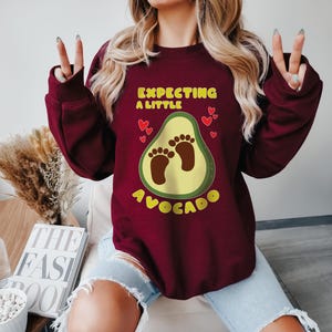May include: A maroon sweatshirt with the text "EXPECTING A LITTLE AVOCADO" in yellow. The design features an avocado with baby footprints and small red hearts. The sweatshirt is a casual style.