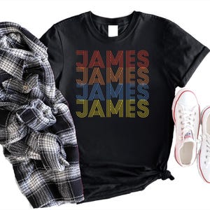 Puede incluir: Camiseta negra con el nombre "JAMES" en letras retro en naranja, rojo, azul y amarillo. Una camisa a cuadros en blanco y negro y zapatillas blancas con ribete rojo tambi&eacute;n est&aacute;n en la imagen.