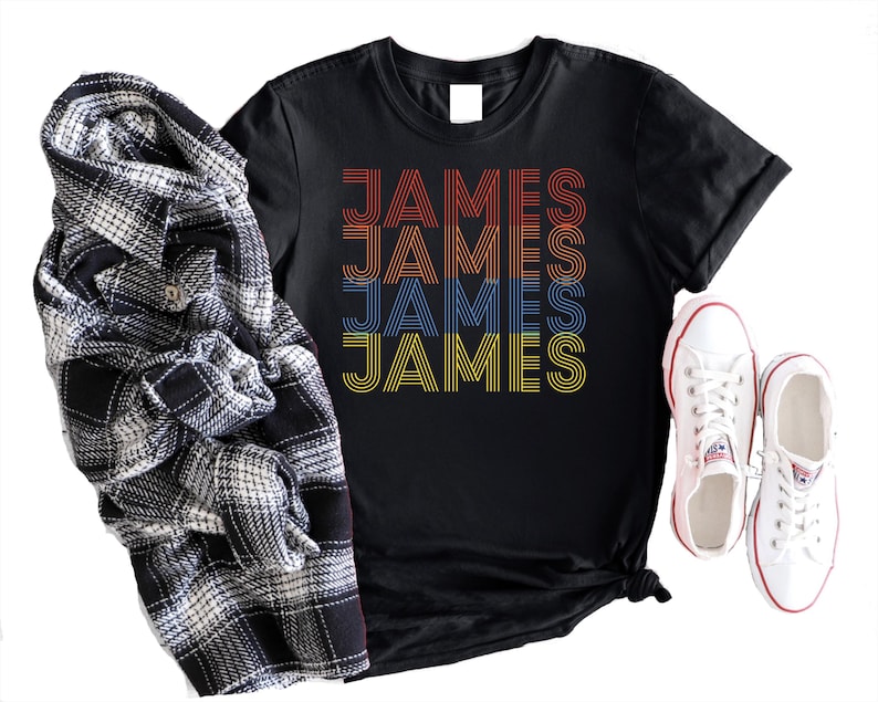 Puede incluir: Camiseta negra con el nombre "JAMES" en letras retro en naranja, rojo, azul y amarillo. Una camisa a cuadros en blanco y negro y zapatillas blancas con ribete rojo tambi&eacute;n est&aacute;n en la imagen.