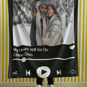 May include: A black and white blanket featuring a couple in a snowy scene. The blanket has a music player interface design with the song title "My Heart Will Go On" by Céline Dion. The blanket is held up by a person wearing black pants and white sneakers.