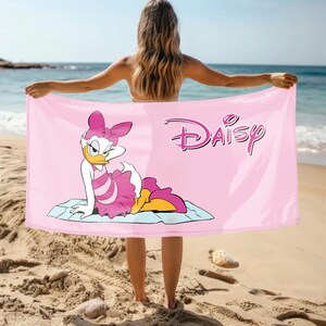 K&ouml;nnte beinhalten: Rosa Strandtuch mit einem Cartoon-Bild von Daisy Duck, die ein rosa Kleid und eine Schleife tr&auml;gt. Das Handtuch hat das Wort "Daisy" in einer stilisierten Schrift gedruckt.