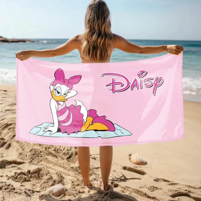 K&ouml;nnte beinhalten: Rosa Strandtuch mit einem Cartoon-Bild von Daisy Duck, die ein rosa Kleid und eine Schleife tr&auml;gt. Das Handtuch hat das Wort "Daisy" in einer stilisierten Schrift gedruckt.