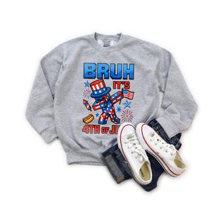 Puede incluir: Una sudadera gris jaspeada con la frase "BRUH IT'S 4TH OF JULY". El dise&ntilde;o presenta un personaje pixelado en rojo, blanco y azul con un sombrero de copa y una bandera americana. Zapatillas blancas y vaqueros tambi&eacute;n en la imagen.