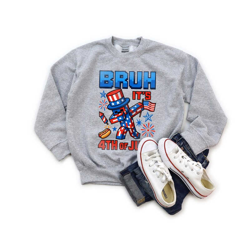 Puede incluir: Una sudadera gris jaspeada con la frase "BRUH IT'S 4TH OF JULY". El dise&ntilde;o presenta un personaje pixelado en rojo, blanco y azul con un sombrero de copa y una bandera americana. Zapatillas blancas y vaqueros tambi&eacute;n en la imagen.