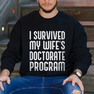 May include: Black sweatshirt with white text that reads "I SURVIVED MY WIFE'S DOCTORATE PROGRAM." The sweatshirt is worn by a person sitting on steps. The person is wearing blue jeans and a watch.
