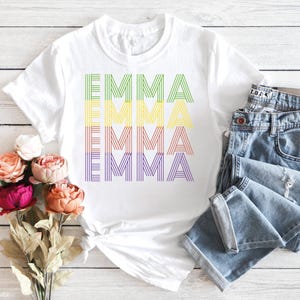 Puede incluir: Camiseta blanca con el nombre "EMMA" impreso en colores del arco&iacute;ris. La palabra est&aacute; apilada verticalmente, con cada letra formada por l&iacute;neas paralelas. Unos vaqueros azules est&aacute;n doblados junto a la camiseta.
