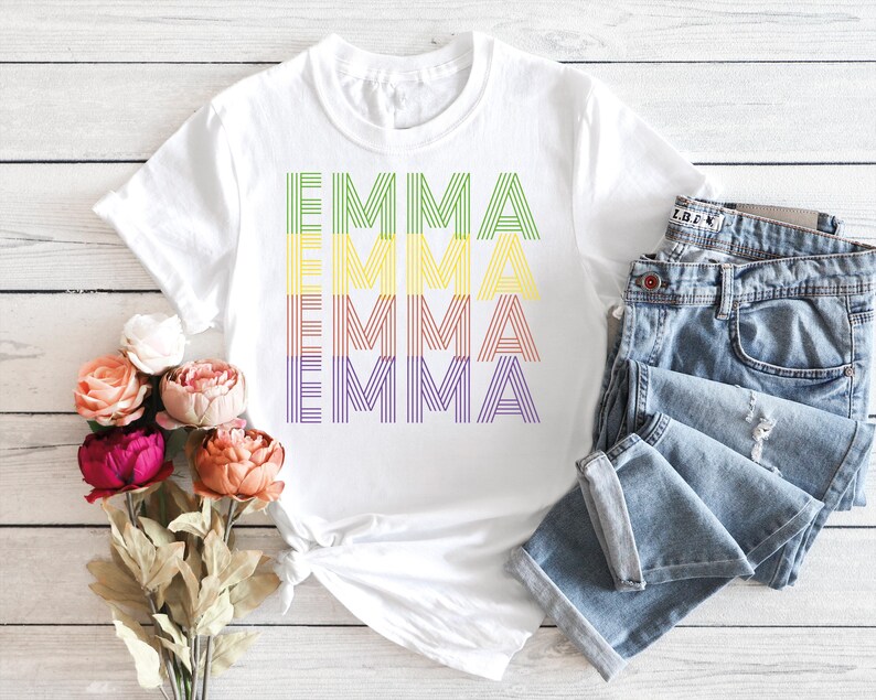 Puede incluir: Camiseta blanca con el nombre "EMMA" impreso en colores del arco&iacute;ris. La palabra est&aacute; apilada verticalmente, con cada letra formada por l&iacute;neas paralelas. Unos vaqueros azules est&aacute;n doblados junto a la camiseta.