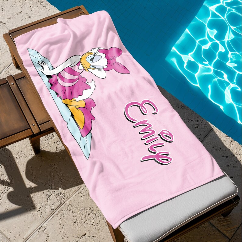 K&ouml;nnte beinhalten: Rosa Strandtuch mit einem Cartoon-Bild eines Entchens, das ein rosa Kleid und eine Schleife tr&auml;gt. Das Handtuch tr&auml;gt den Namen "Emily" in rosa Kursivschrift.