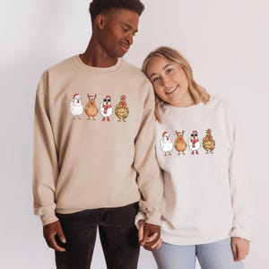 May include: Two people wearing matching beige and off-white sweatshirts. Each sweatshirt features a whimsical illustration of four anthropomorphic chickens dressed in holiday attire, including Santa hats, reindeer antlers, and sunglasses. The sweatshirts are a festive and fun holiday theme.