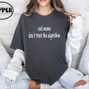 May include: Dark gray t-shirt with the text "cool moms don't trust the algorithm" in white. The shirt is a relaxed fit and is worn with light-wash jeans and a gray sweater. The image includes a Pepper logo in a black oval.