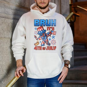 Puede incluir: Sudadera con capucha blanca con un gr&aacute;fico que dice "BRUH IT'S 4TH OF JULY". El gr&aacute;fico presenta un personaje patri&oacute;tico con un sombrero de copa, sosteniendo una bandera y un perrito caliente, con fuegos artificiales y estrellas.
