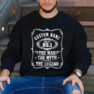 May include: Black sweatshirt with white text that reads "CUSTOM NAME BEST NO.1 THE MAN THE MYTH THE LEGEND". The sweatshirt is worn by a person sitting on wooden steps, wearing blue jeans and a watch.