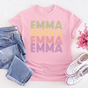Puede incluir: Una camiseta rosa con el nombre "EMMA" impreso en un dise&ntilde;o a rayas en verde, amarillo, naranja y morado. La imagen incluye vaqueros azules, zapatillas blancas y un ramo de flores rosas.