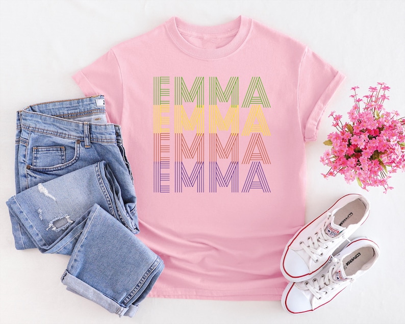 Puede incluir: Una camiseta rosa con el nombre "EMMA" impreso en un dise&ntilde;o a rayas en verde, amarillo, naranja y morado. La imagen incluye vaqueros azules, zapatillas blancas y un ramo de flores rosas.