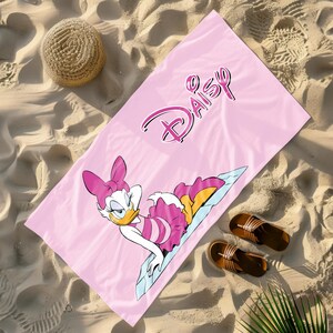 K&ouml;nnte beinhalten: Rosa Strandtuch mit einem Cartoon-Bild von Daisy Duck, die einen rosa-wei&szlig;en Badeanzug tr&auml;gt. Das Handtuch tr&auml;gt den Schriftzug "Daisy" in rosa kursiver Schrift.