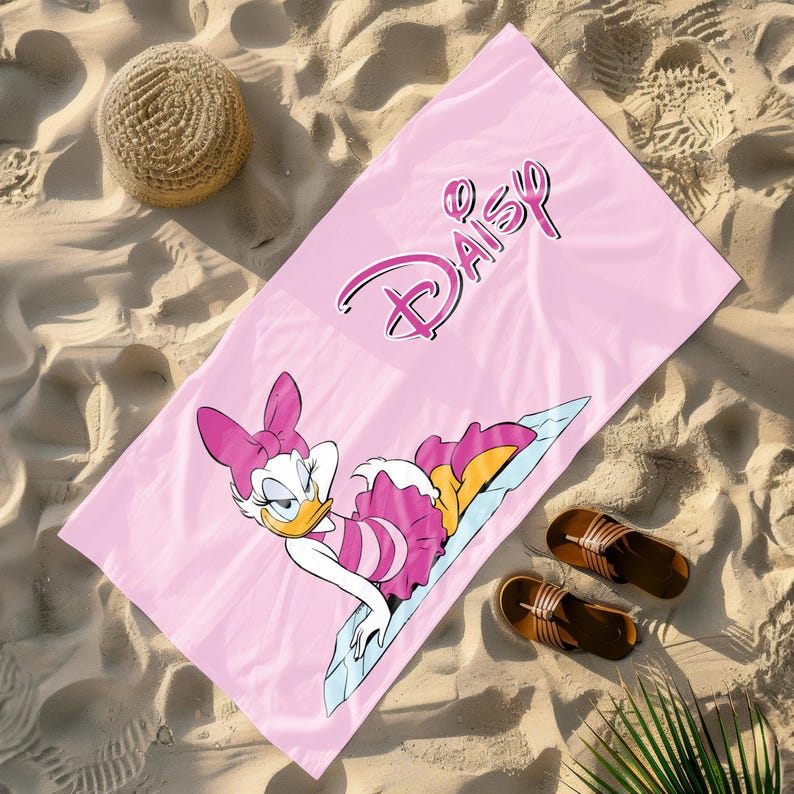 K&ouml;nnte beinhalten: Rosa Strandtuch mit einem Cartoon-Bild von Daisy Duck, die einen rosa-wei&szlig;en Badeanzug tr&auml;gt. Das Handtuch tr&auml;gt den Schriftzug "Daisy" in rosa kursiver Schrift.