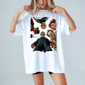 May include: White t-shirt featuring a graphic design of horror movie characters, including Michael Myers, Freddy Krueger, Jason Voorhees, Chucky, and Pennywise. The design is in a cartoon style with vibrant colors.