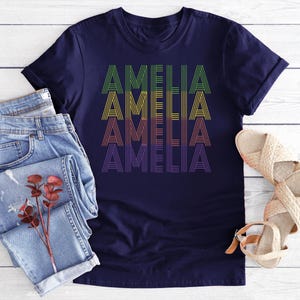 Puede incluir: Una camiseta azul marino con el nombre "AMELIA" impreso en un dise&ntilde;o apilado. El texto presenta una gradaci&oacute;n de colores, de verde a morado. La camiseta se muestra con vaqueros azules claros y sandalias tejidas.