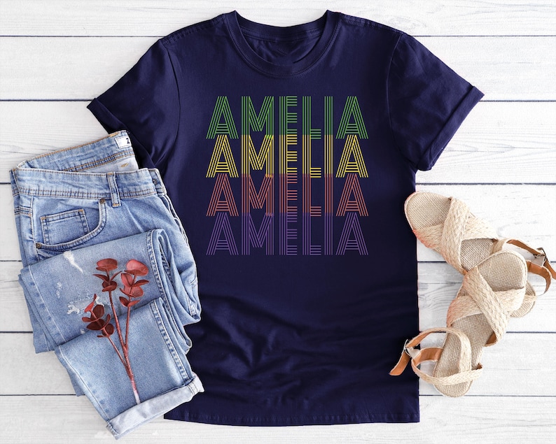 Puede incluir: Una camiseta azul marino con el nombre "AMELIA" impreso en un dise&ntilde;o apilado. El texto presenta una gradaci&oacute;n de colores, de verde a morado. La camiseta se muestra con vaqueros azules claros y sandalias tejidas.