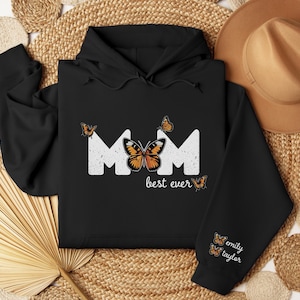 May include: Black hoodie with the word "MOM" in white, featuring butterfly graphics. The phrase "best ever" is below the word "MOM". The sleeve has the names "emily taylor" and butterfly graphics.
