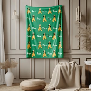 May include: A green blanket featuring a repeating pattern of a cartoon dog with yellow fur and black ears. The word "Pluto" is written in white script above each dog. The blanket is displayed on a wall.