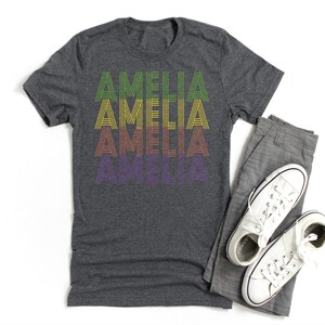 Puede incluir: Una camiseta gris oscuro con el nombre "AMELIA" impreso en estilo retro en verde, amarillo, naranja y morado. La camiseta se muestra con pantalones grises y zapatillas blancas.