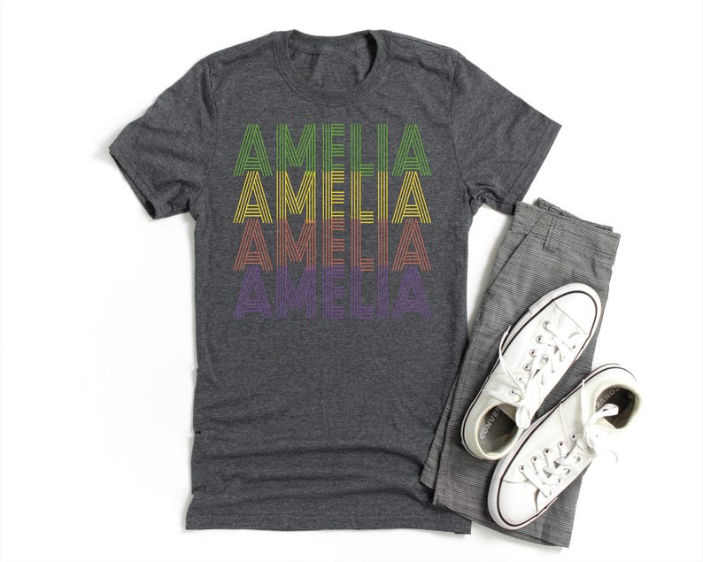 Puede incluir: Una camiseta gris oscuro con el nombre "AMELIA" impreso en estilo retro en verde, amarillo, naranja y morado. La camiseta se muestra con pantalones grises y zapatillas blancas.