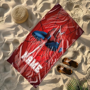May include: Red beach towel featuring Spider-Man in a web design, with the word "NAME" in white block letters. The towel is on a sandy beach, with a straw hat and sandals nearby. The towel is rectangular.