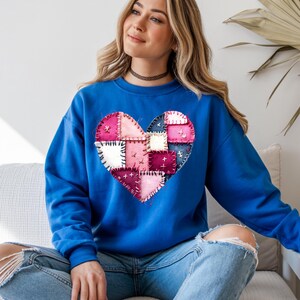May include: A royal blue sweatshirt featuring a patchwork heart design in shades of pink, white, and burgundy. The heart is composed of various fabric squares stitched together, creating a textured effect. The sweatshirt has long sleeves and a crew neck.
