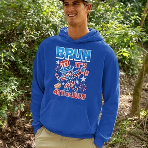 Puede incluir: Una sudadera con capucha azul real con un gr&aacute;fico que dice "BRUH IT'S 4TH OF JULY". El gr&aacute;fico presenta una figura patri&oacute;tica del T&iacute;o Sam, fuegos artificiales y un perrito caliente. La persona de la imagen lleva una gorra verde y pantalones color caqui.