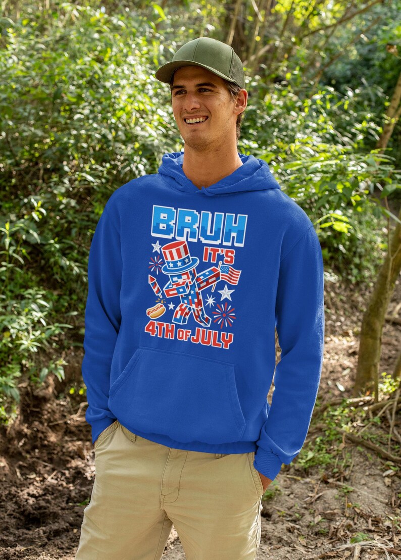 Puede incluir: Una sudadera con capucha azul real con un gr&aacute;fico que dice "BRUH IT'S 4TH OF JULY". El gr&aacute;fico presenta una figura patri&oacute;tica del T&iacute;o Sam, fuegos artificiales y un perrito caliente. La persona de la imagen lleva una gorra verde y pantalones color caqui.