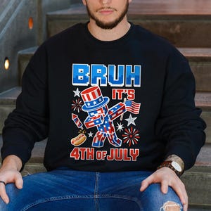 Puede incluir: Sudadera negra con un gr&aacute;fico patri&oacute;tico. El dise&ntilde;o presenta una figura pixelada con sombrero de copa, sosteniendo una bandera, con el texto "BRUH IT'S 4TH OF JULY" en rojo y azul. Tambi&eacute;n se incluye un perrito caliente y fuegos artificiales.