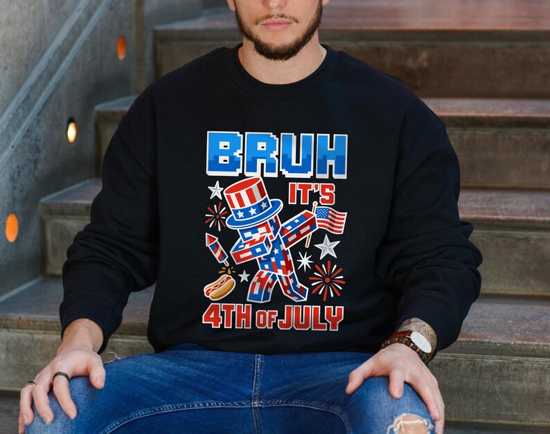 Puede incluir: Sudadera negra con un gr&aacute;fico patri&oacute;tico. El dise&ntilde;o presenta una figura pixelada con sombrero de copa, sosteniendo una bandera, con el texto "BRUH IT'S 4TH OF JULY" en rojo y azul. Tambi&eacute;n se incluye un perrito caliente y fuegos artificiales.