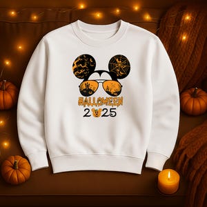 May include: White sweatshirt with a Halloween-themed graphic. The design features Mickey Mouse ears with bats and a spiderweb, sunglasses with a haunted house scene, and the text "HALLOWEEN 2025" with a pumpkin.