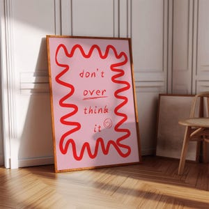 May include: A framed pink art print with red wavy lines and the words "don't over think it" in red. A small smiley face is below the text. The frame is brown and the background is a light pink.
